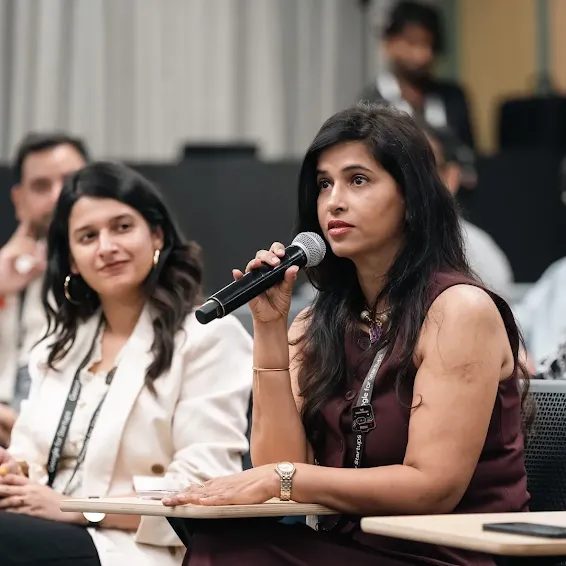 A woman speaking into a microphone while another woman listens seated beside her at an event.