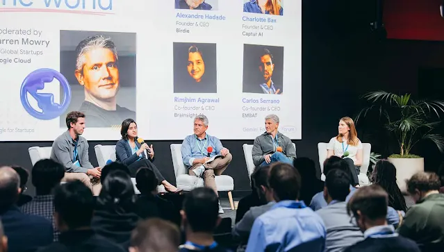 Five panelists sitting in armchairs on a stage during a panel discussion, with one woman speaking into a microphone. A large presentation screen behind them displays speaker profiles, and an audience watches from the foreground.